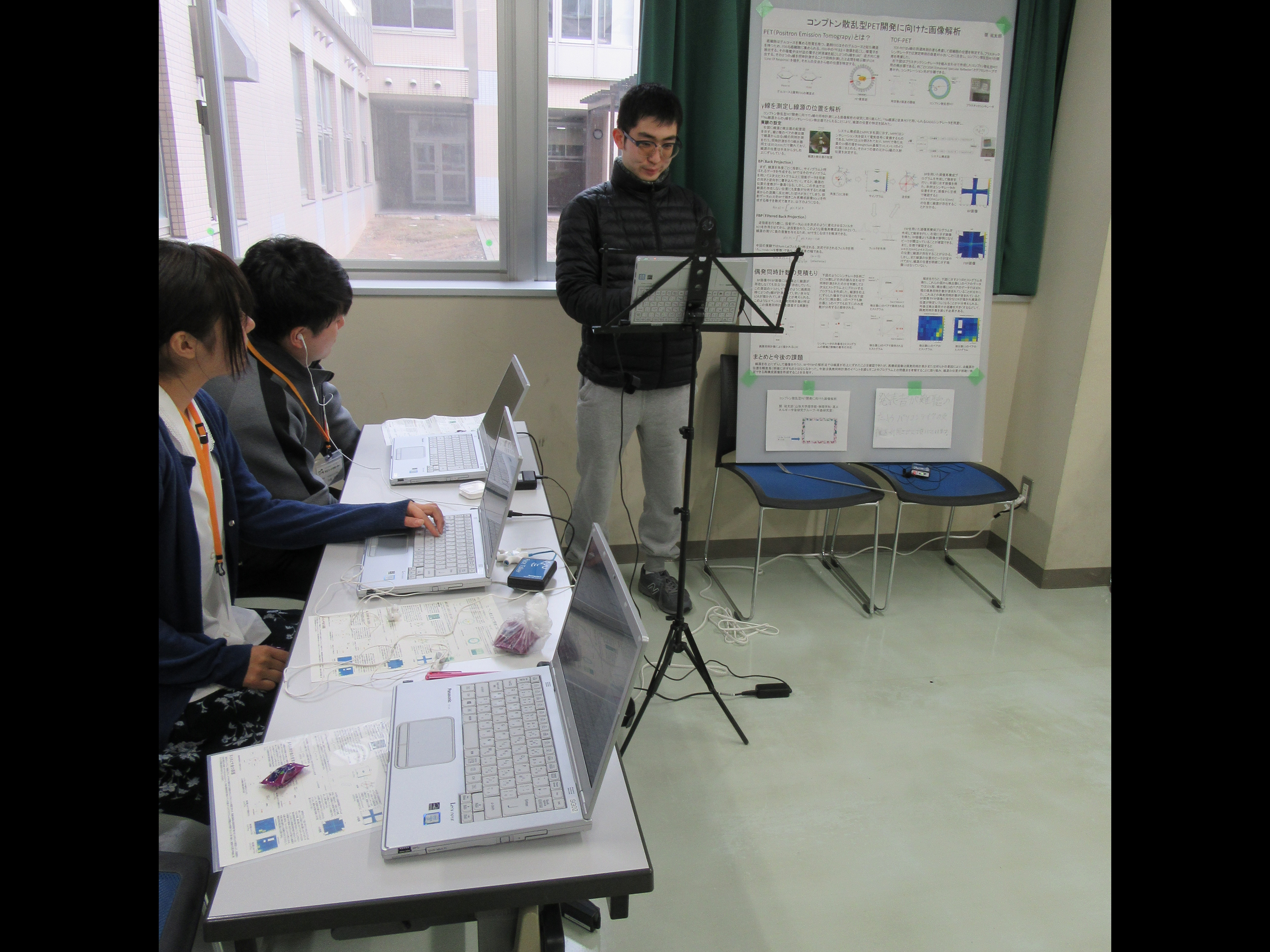 真ん中に聴覚障がい学生が立っておりその左隣に支援学生が２名いる。支援学生の前にはパソコンが置かれている。