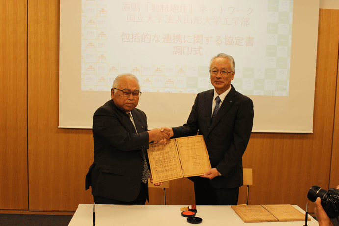 木製の協定書を挟んで握手を交わす河内会長（左）と飯塚工学部長の画像