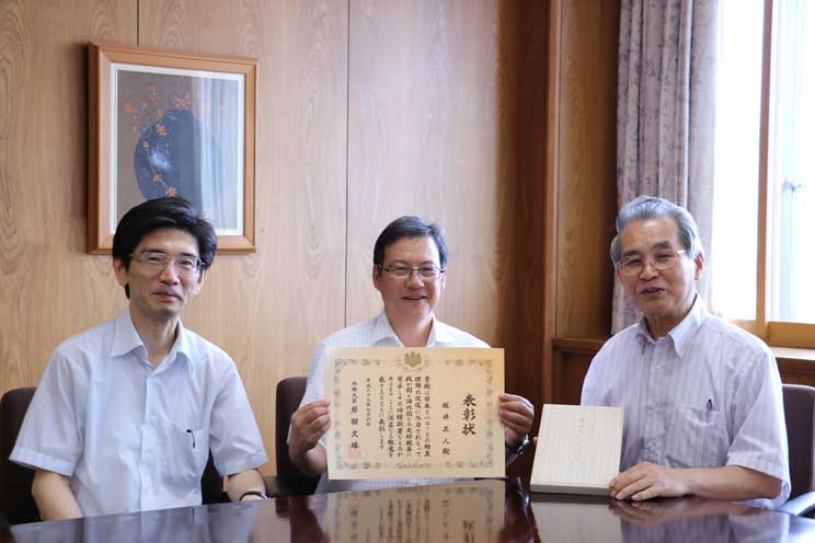 賞状を手に喜びを報告する坂井教授（中央）、清塚人文社会科学部長（左）、副賞を手にする学長（右)の画像